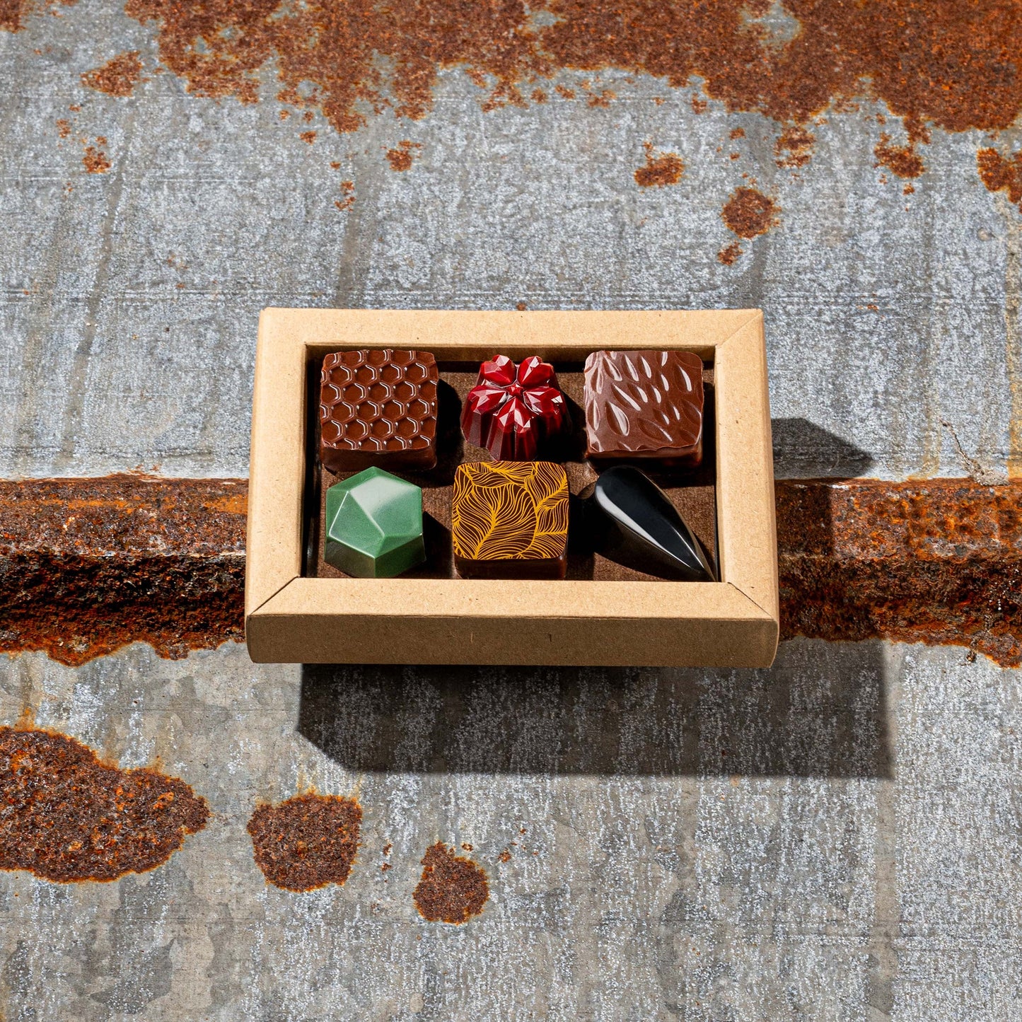 La Feve chocolate box  with 6 flavours bonbons. 3 square pralines and 3 jewel shaped bonbons on a rustic metal surface