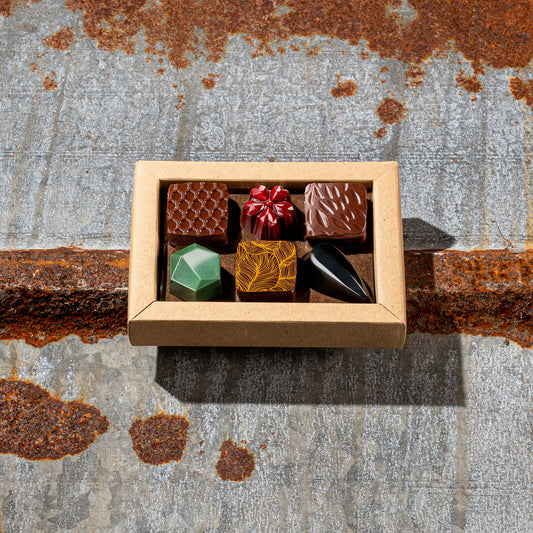 La Feve chocolate box  with 6 flavours bonbons. 3 square pralines and 3 jewel shaped bonbons on a rustic metal surface